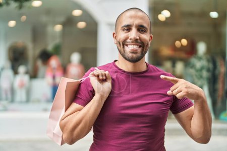 Téléchargez les photos : Homme hispanique avec barbe tenant des sacs à provisions à l'extérieur souriant heureux pointant avec la main et le doigt - en image libre de droit