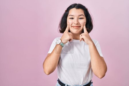 Téléchargez les photos : Jeune femme hispanique portant un t-shirt blanc décontracté sur fond rose souriant avec bouche ouverte, doigts pointant et forçant sourire joyeux - en image libre de droit