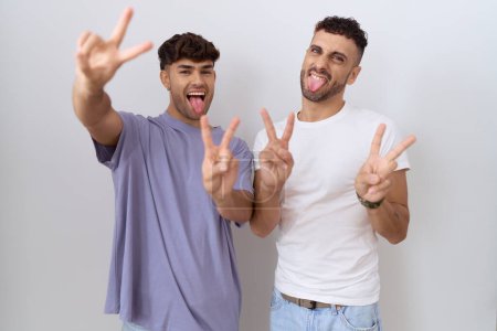 Téléchargez les photos : Couple homosexuel gay debout sur fond blanc souriant avec la langue montrant les doigts des deux mains faisant signe de victoire. numéro deux. - en image libre de droit
