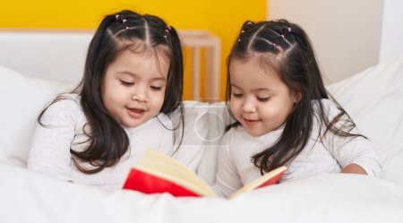 Photo for Adorable twin girls reading book sitting on bed at bedroom - Royalty Free Image