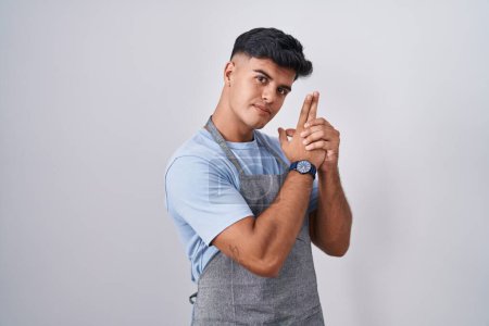 Photo for Hispanic young man wearing apron over white background holding symbolic gun with hand gesture, playing killing shooting weapons, angry face - Royalty Free Image