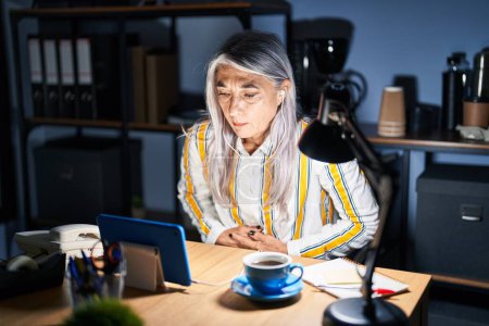 Photo for Middle age woman with grey hair working at the office at night with hand on stomach because indigestion, painful illness feeling unwell. ache concept. - Royalty Free Image