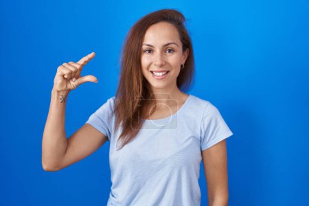 Téléchargez les photos : Femme brune debout sur fond bleu geste souriant et confiant avec la main faisant signe de petite taille avec les doigts regardant et la caméra. concept de mesure. - en image libre de droit