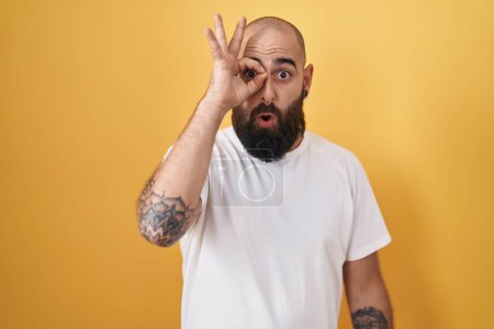 Foto de Joven hombre hispano con barba y tatuajes de pie sobre fondo amarillo haciendo un gesto bien sorprendido con la cara sorprendida, los ojos mirando a través de los dedos. expresión incrédula. - Imagen libre de derechos