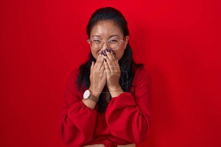 Foto de Asiática joven mujer de pie sobre rojo fondo riendo y avergonzado risita cubriendo la boca con las manos, chismes y escándalo concepto - Imagen libre de derechos