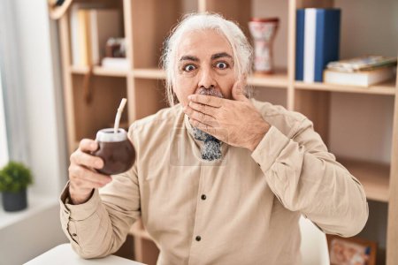 Photo for Middle age man with grey hair drinking mate infusion covering mouth with hand, shocked and afraid for mistake. surprised expression - Royalty Free Image