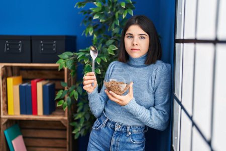 Photo for Young hispanic woman eating healthy whole grain cereals with spoon relaxed with serious expression on face. simple and natural looking at the camera. - Royalty Free Image