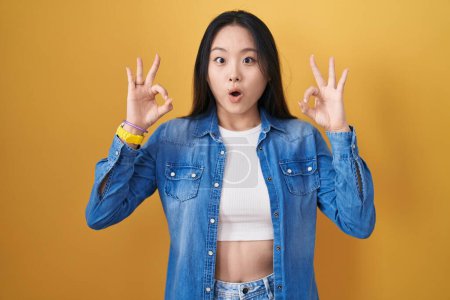 Téléchargez les photos : Jeune femme asiatique debout sur fond jaune regardant surpris et choqué faire ok symbole d'approbation avec les doigts. expression folle - en image libre de droit