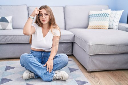 Photo for Young caucasian woman sitting on the floor at the living room pointing unhappy to pimple on forehead, ugly infection of blackhead. acne and skin problem - Royalty Free Image