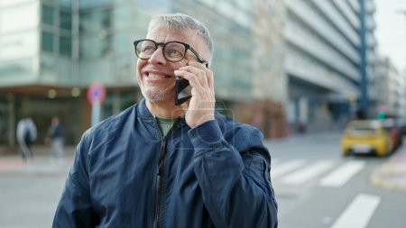 Téléchargez les photos : Moyen âge homme aux cheveux gris souriant confiant parler sur smartphone à la rue - en image libre de droit