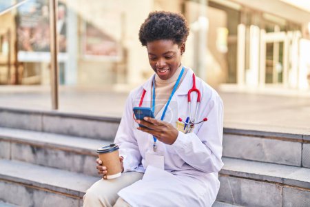 Téléchargez les photos : Femme afro-américaine portant l'uniforme de médecin en utilisant smartphone boire du café dans la rue - en image libre de droit