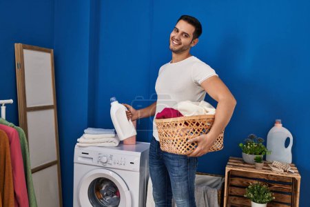 Photo for Young hispanic man holding basket with clothes and detergent bottle at laundry room - Royalty Free Image