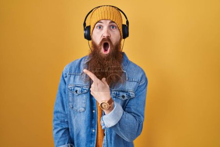 Foto de Hombre caucásico con barba larga escuchando música usando auriculares sorprendidos apuntando con el dedo hacia un lado, boca abierta expresión asombrada. - Imagen libre de derechos