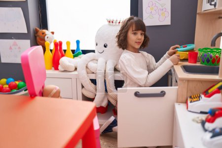 Photo for Adorable hispanic girl playing with play kitchen sitting on floor at kindergarten - Royalty Free Image