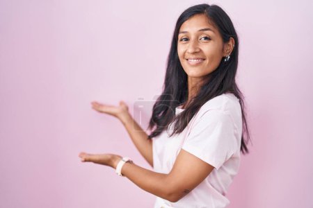 Photo for Young hispanic woman standing over pink background inviting to enter smiling natural with open hand - Royalty Free Image