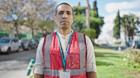 Téléchargez les photos : Jeune homme hispanique bénévole portant un gilet regardant sérieusement au parc - en image libre de droit