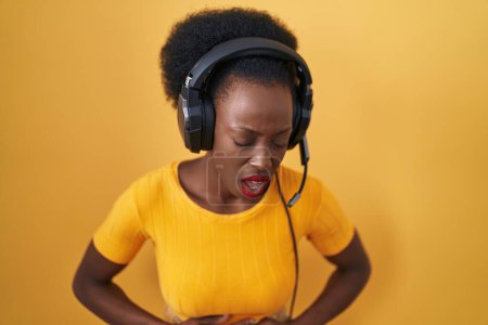 Téléchargez les photos : Femme africaine avec les cheveux bouclés debout sur fond jaune portant des écouteurs avec la main sur le ventre parce que l'indigestion, la maladie douloureuse se sent mal. concept de douleur. - en image libre de droit