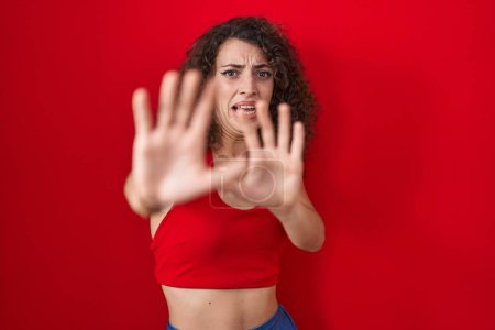 Téléchargez les photos : Femme hispanique avec les cheveux bouclés debout sur fond rouge peur et terrifié par l'expression de la peur arrêter geste avec les mains, criant en état de choc. concept de panique. - en image libre de droit