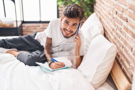 Foto de Joven hispano hablando en un smartphone escribiendo en un libro en el dormitorio - Imagen libre de derechos