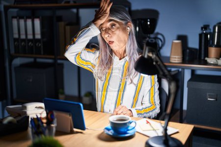 Téléchargez les photos : Femme d'âge moyen avec les cheveux gris travaillant au bureau la nuit surpris avec la main sur la tête pour erreur, rappelez-vous erreur. oublié, mauvais concept de mémoire. - en image libre de droit