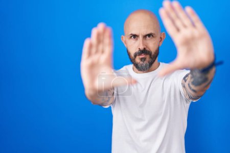 Foto de Hombre hispano con tatuajes de pie sobre fondo azul haciendo marco usando manos palmas y dedos, perspectiva de la cámara - Imagen libre de derechos