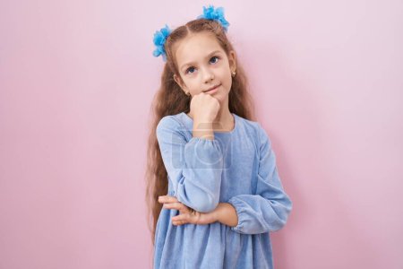 Photo for Young little girl standing over pink background serious face thinking about question with hand on chin, thoughtful about confusing idea - Royalty Free Image