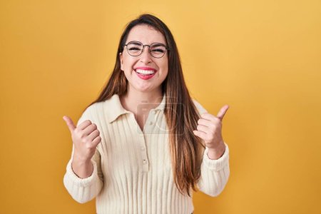 Foto de Young hispanic woman standing over yellow background success sign doing positive gesture with hand, thumbs up smiling and happy. cheerful expression and winner gesture. - Imagen libre de derechos