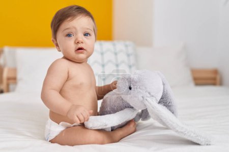 Foto de Adorable bebé caucásico sentado en la cama jugando con la muñeca en el dormitorio - Imagen libre de derechos