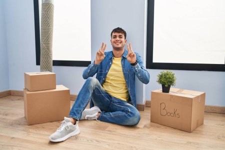 Photo for Young hispanic man sitting on the floor at new home smiling looking to the camera showing fingers doing victory sign. number two. - Royalty Free Image