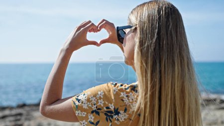 Téléchargez les photos : Jeune femme blonde faisant un geste cardiaque vers l'arrière au bord de la mer - en image libre de droit