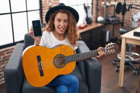 Photo for Young caucasian woman playing classic guitar at music studio holding smartphone sticking tongue out happy with funny expression. - Royalty Free Image