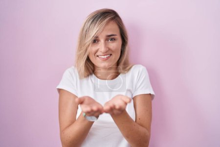 Téléchargez les photos : Jeune femme blonde debout sur fond rose souriant avec les mains paumes ensemble recevant ou donnant geste. maintien et protection - en image libre de droit