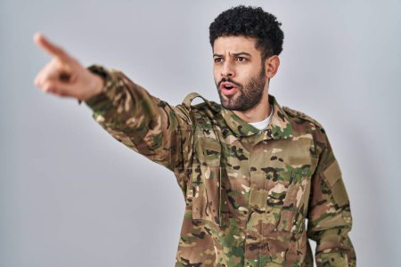 Photo for Arab man wearing camouflage army uniform pointing with finger surprised ahead, open mouth amazed expression, something on the front - Royalty Free Image