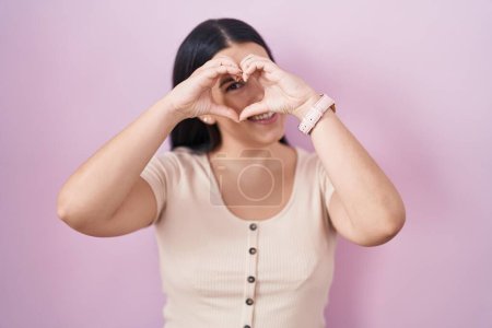 Photo for Young hispanic woman standing over pink background doing heart shape with hand and fingers smiling looking through sign - Royalty Free Image