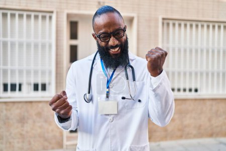 Téléchargez les photos : Homme afro-américain portant l'uniforme de médecin et stéthoscope célébrant surpris et étonnés pour le succès avec les bras levés et les yeux fermés - en image libre de droit