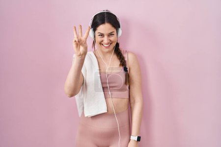 Photo for Young brunette woman wearing sportswear and headphones showing and pointing up with fingers number three while smiling confident and happy. - Royalty Free Image