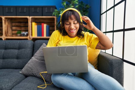 Foto de Young beautiful latin woman using laptop and headphones sitting on sofa at home - Imagen libre de derechos