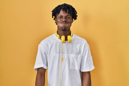 Téléchargez les photos : Jeune homme africain avec dreadlocks debout sur fond jaune joues gonflées avec drôle de visage. bouche gonflée d'air, expression folle. - en image libre de droit