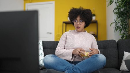 Photo for Young african american woman watching tv sitting on sofa eating chips at home - Royalty Free Image
