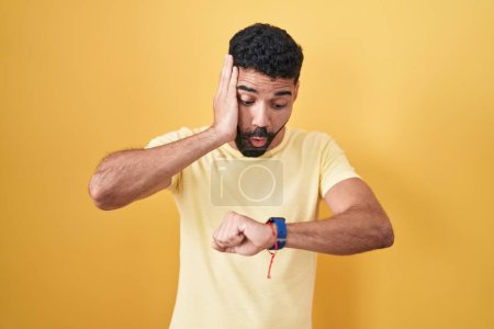 Téléchargez les photos : Homme hispanique avec barbe debout sur fond jaune regardant le temps de la montre inquiet, peur de se faire tard - en image libre de droit