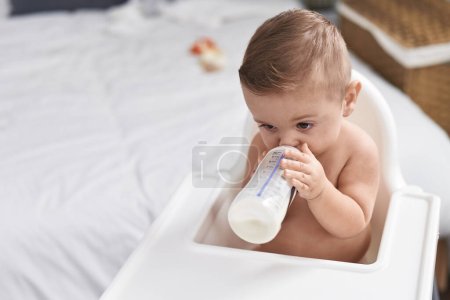 Téléchargez les photos : Adorable bébé caucasien sucer du lait sur le biberon assis sur chaise haute à la chambre - en image libre de droit
