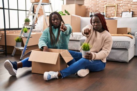 Téléchargez les photos : Deux femmes africaines se déplaçant vers une nouvelle maison assise sur le sol pointant du doigt la caméra et à vous, geste confiant regardant sérieux - en image libre de droit