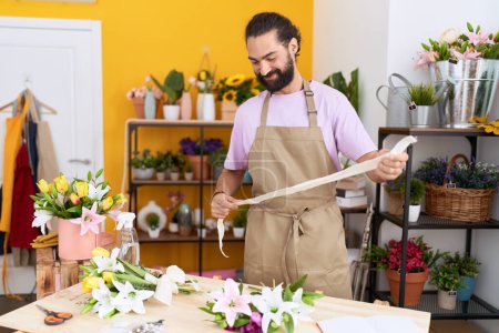 Téléchargez les photos : Jeune homme hispanique fleuriste souriant confiant tenant dentelle cadeau à la boutique de fleurs - en image libre de droit
