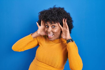 Téléchargez les photos : Femme noire avec les cheveux bouclés debout sur fond bleu souriant joyeux jeu jeter un coup d'oeil avec les mains montrant le visage. surpris et sortis - en image libre de droit