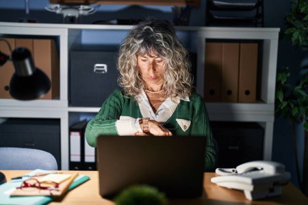 Téléchargez les photos : Femme d'âge moyen travaillant la nuit à l'aide d'un ordinateur portable vérifiant l'heure sur la montre-bracelet, détendu et confiant - en image libre de droit