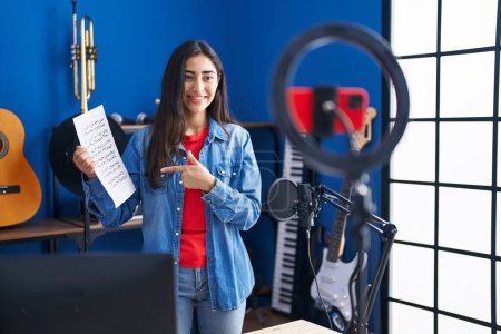 Téléchargez les photos : Jeune adolescente faisant tutoriel de musique en ligne montrant feuille de musique souriant heureux pointant avec la main et le doigt - en image libre de droit