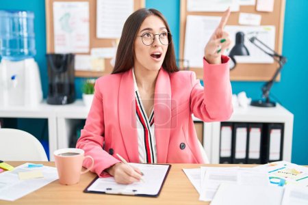Photo for Young hispanic woman working at the office wearing glasses pointing with finger surprised ahead, open mouth amazed expression, something on the front - Royalty Free Image