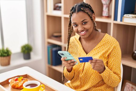 Téléchargez les photos : Femme afro-américaine utilisant smartphone et carte de crédit prendre le petit déjeuner à la maison - en image libre de droit