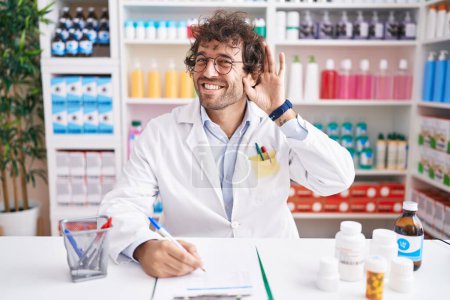 Foto de Joven hispano trabajando en farmacia sonriendo con la mano sobre el oído escuchando un rumor o chismes. concepto de sordera. - Imagen libre de derechos