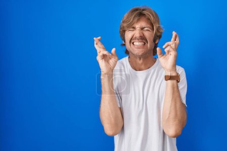 Téléchargez les photos : Homme du Moyen Âge debout sur fond bleu geste doigt croisé souriant avec espoir et les yeux fermés. chance et concept superstitieux. - en image libre de droit